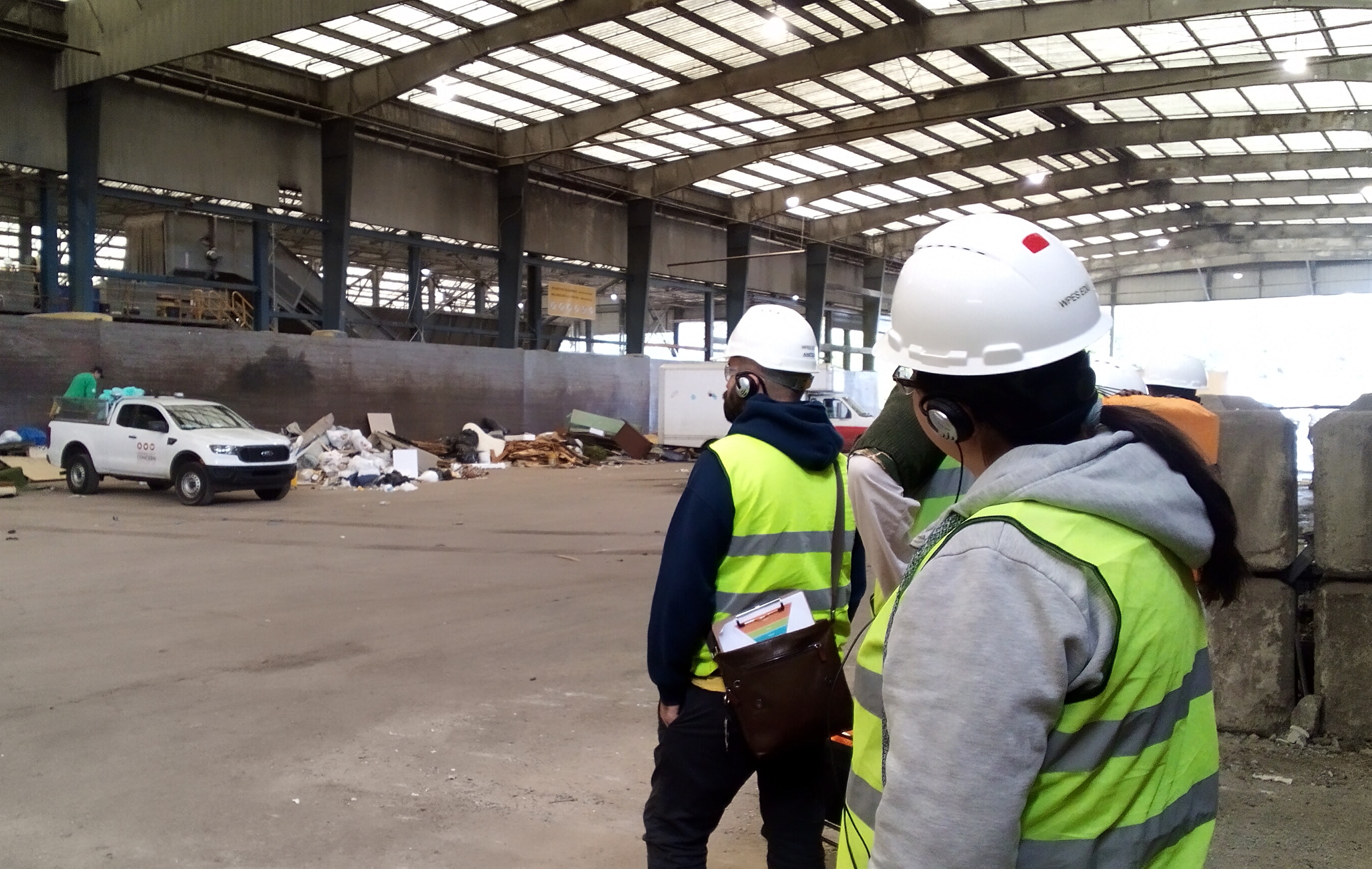 People in bright yellow safety vests and white hard hats stand under a large outdoor covering with vehicles and debris along the far side of the structure.