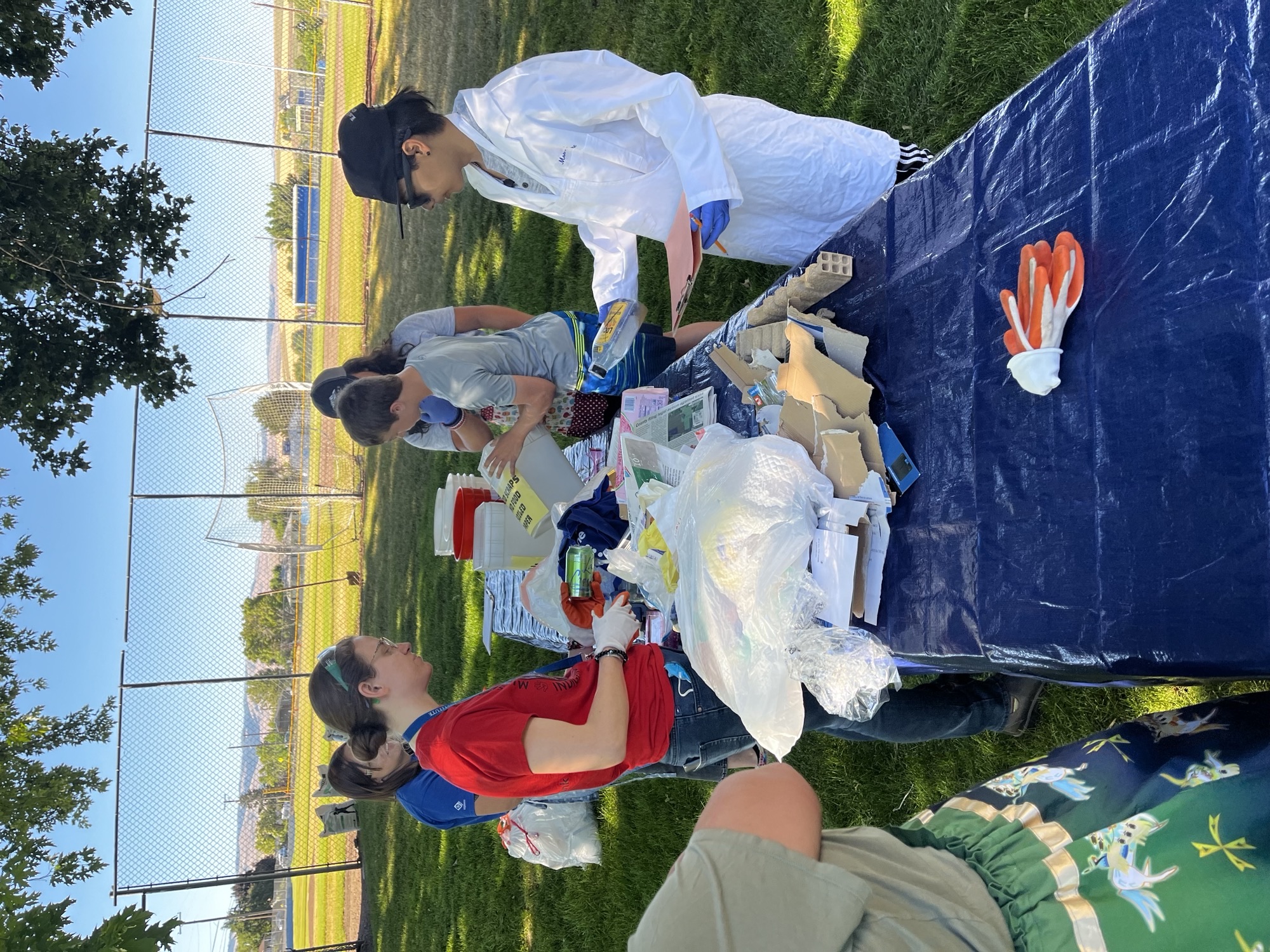 A group of students and teachers gathers around several large black trash bags outside in the shade as they spread the project activity trash out on tables covered with blue tarps.