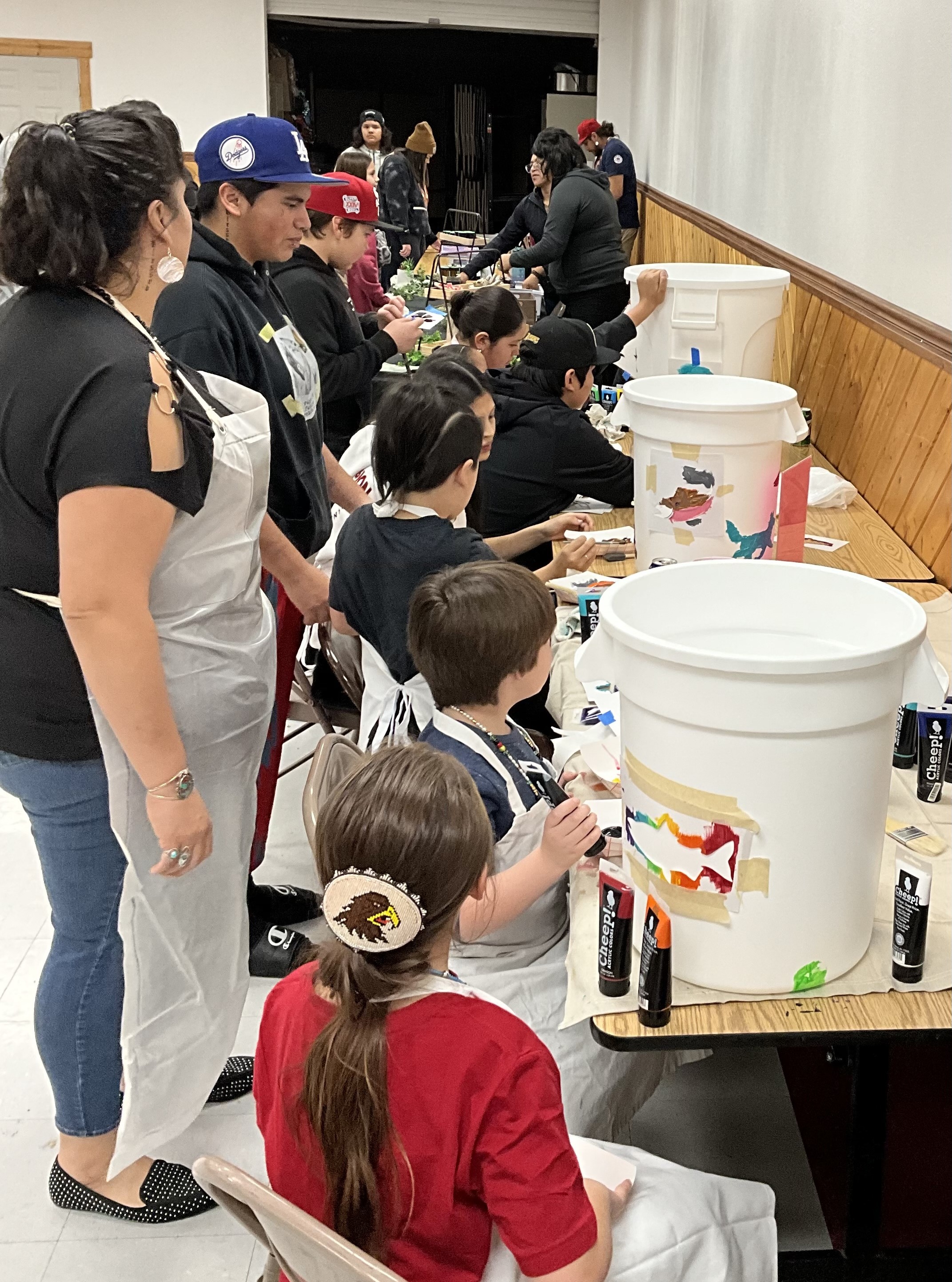 A crowd of people sit or stand at a Longhouse table with multiple white trash bins, as they paint colorful shapes on the bins. Other event vendors with black fabric and products on their table can be seen in the background.