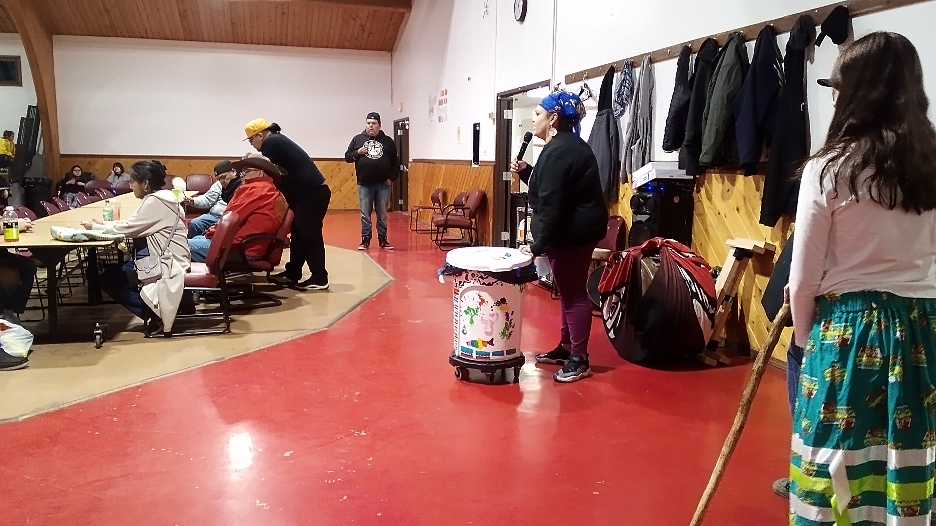 DeArcie stands at the head of the Longhouse main hall speaking into a microphone to the participants of Culture Night. She has a white trash bin on wheels that has been painted colorfully.