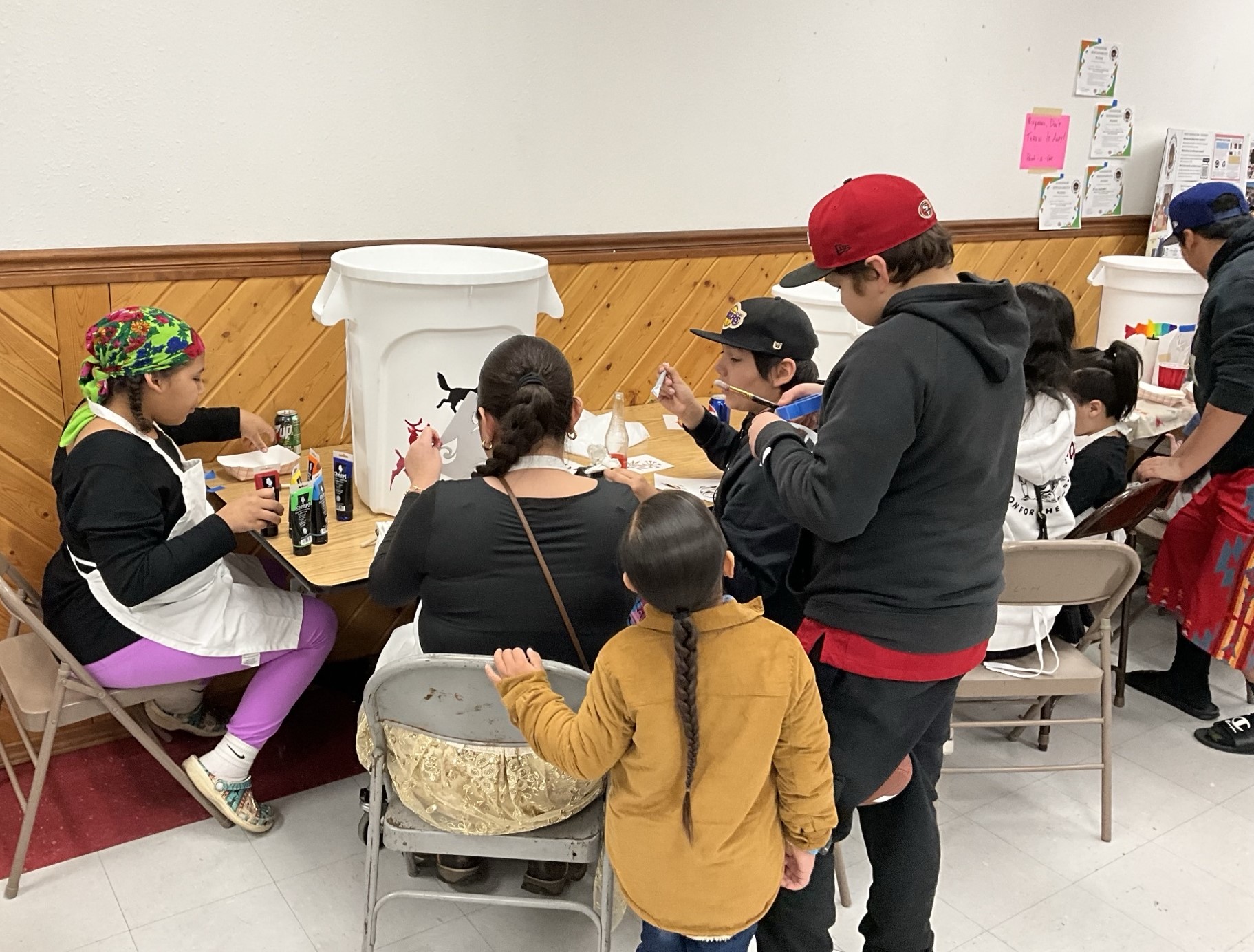 A group of people of different ages sit together at a beige table with a white garbage bin in the middle of them on the table. The people are painting colorful shapes on the white can.