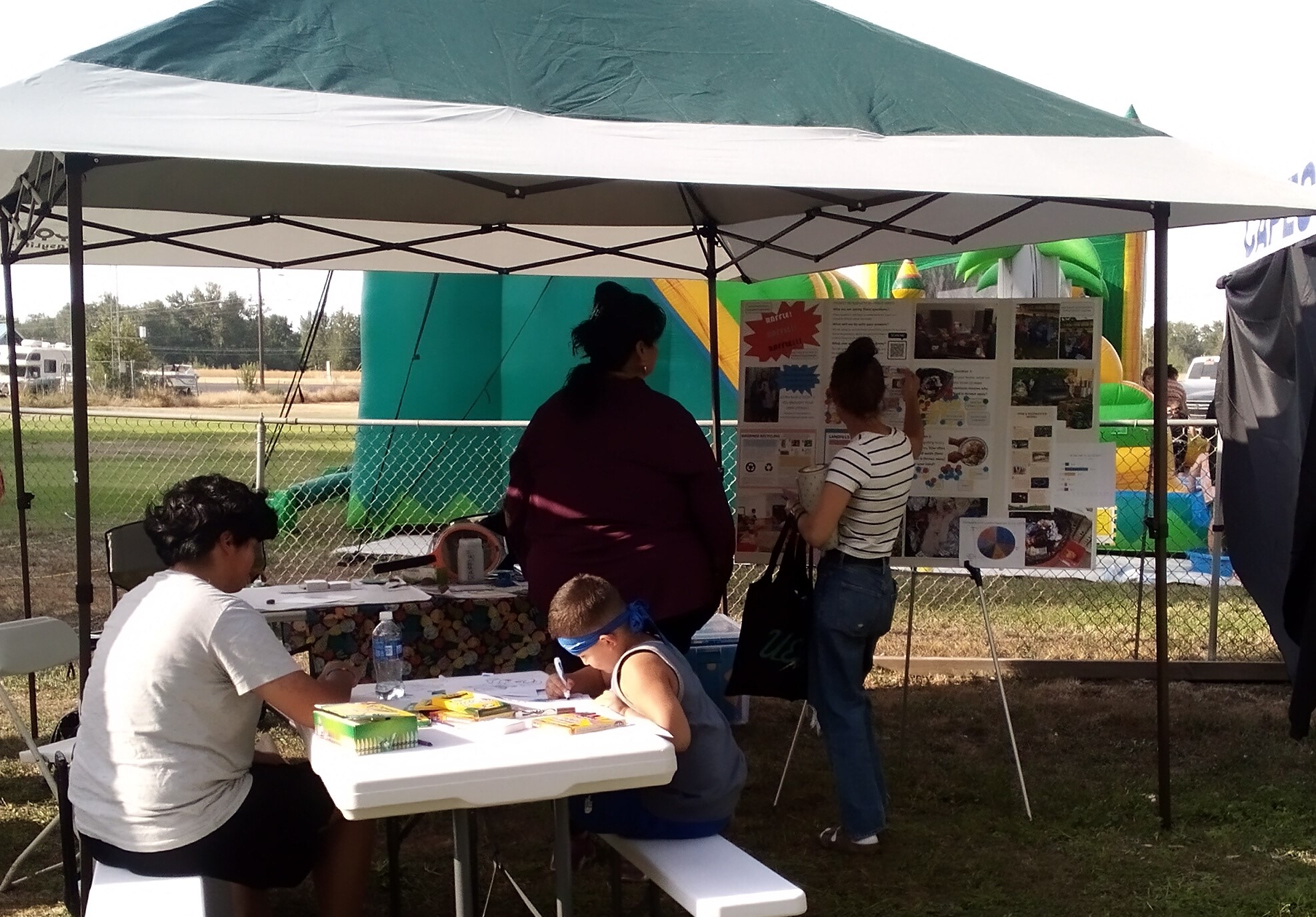 Two people stand and chat next to a poster board with images and text on it under a shade canopy. Two youth sit at a table in front of the canopy and color activity pages on a sunny day.