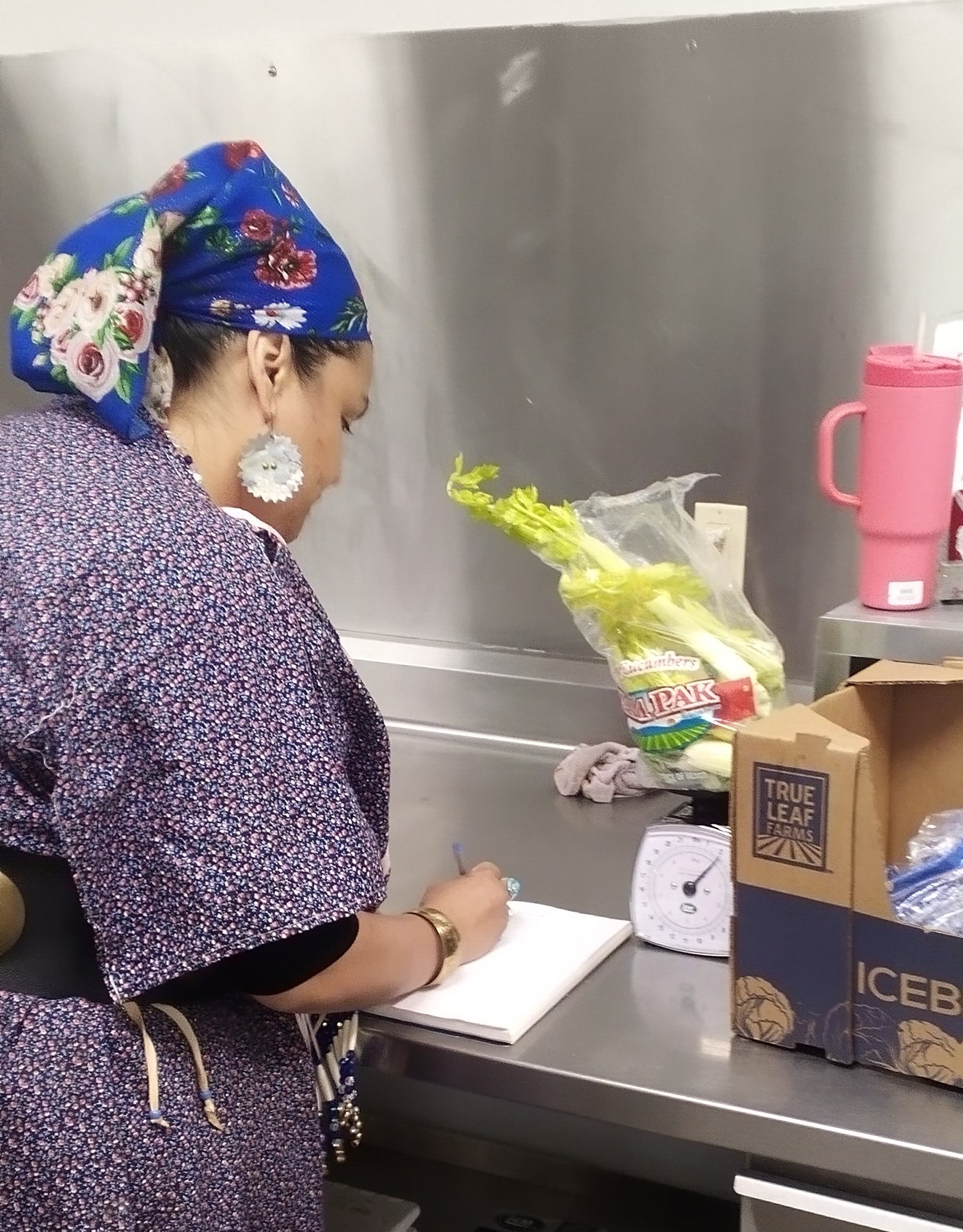 A person in a wing dress and head scarf weighs a bag of food waste on a scale on and writes down the amount on a pad of paper on the counter in the Longhouse kitchen.