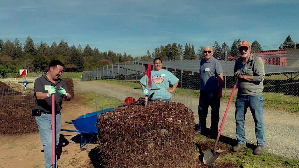 Four people stand smiling around a compost bin