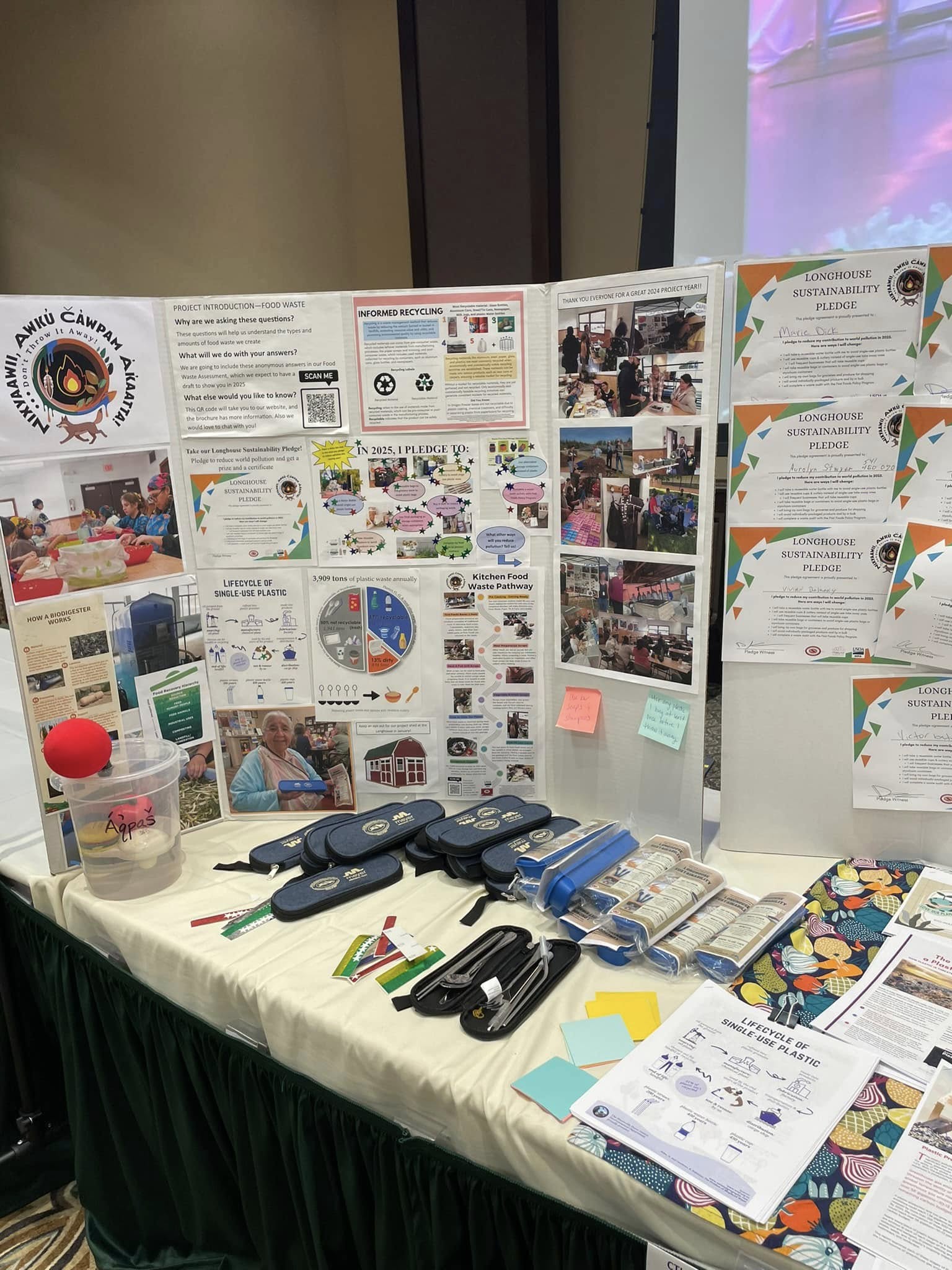 A large cardboard trifold stands at a table in the Wildhorse Rivers Event Center Cayuse Room, covered with colorful infographics, photos, and certificates.