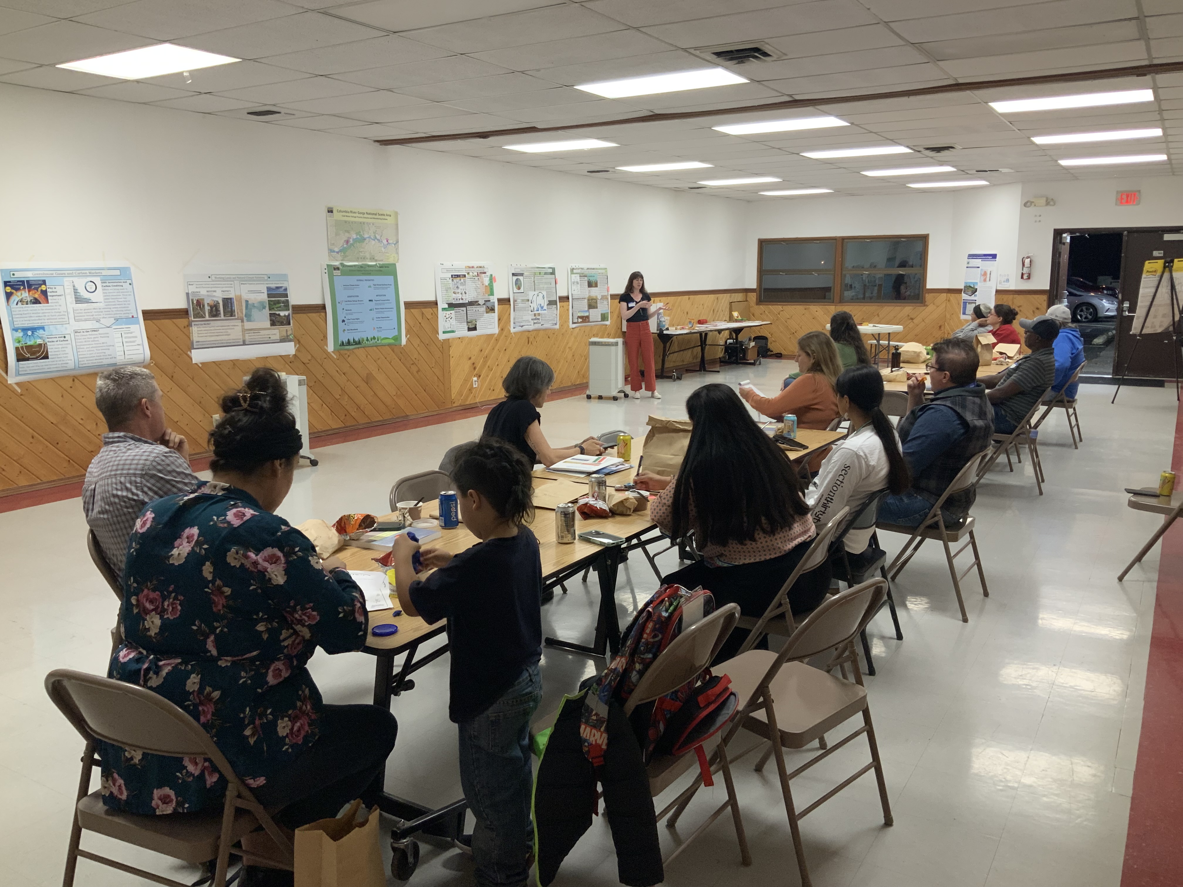 A group of people sit at long tables in the brightly lit Longhouse annex and look at a presenter on the far side of the room, gesturing to one of the many posters taped on the far wall.