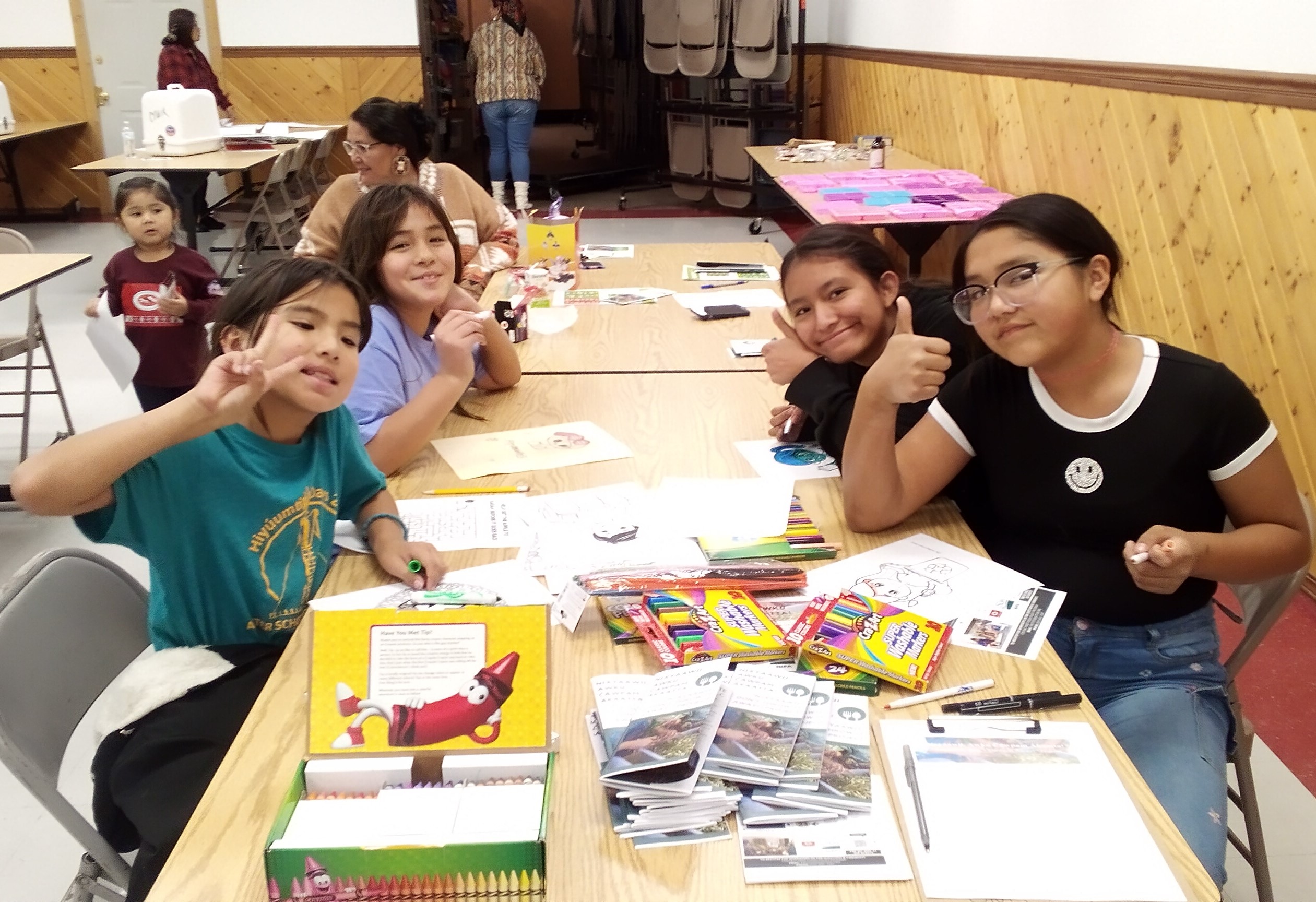 Tribal youth sit at a Longhouse table and smile as they color recycling and composting themed coloring pages at Culture Night. Project pamphlets and educational information can be seen at the front of the table.