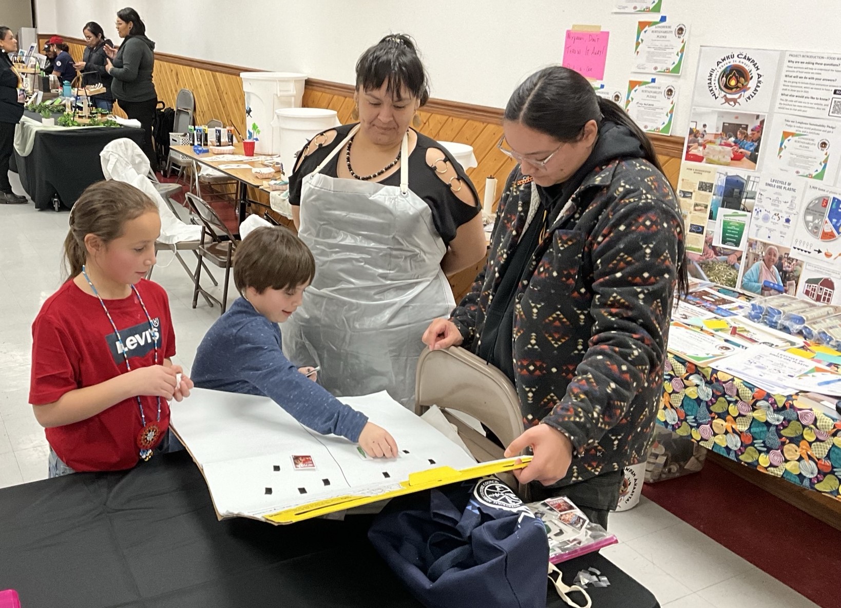 Two adults and two youth stand together and look over a large white tablet with small shapes placed in strategic ways. In the background are other Native vendors at a Longhouse table with a black fabric and products on top.