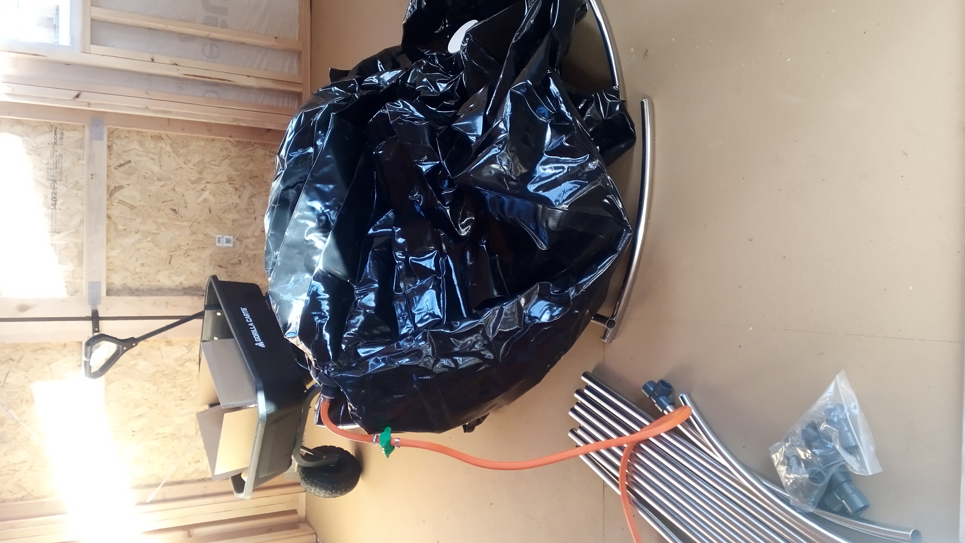 A large black plastic bag sits in the center of the interior of a utility shed with sunlight coming in through windows on all sides of the walls. Metal pipes and other parts are on the floor and shelves of the shed, and a sturdy four-wheeled pull behind cart has a carboard box inside of it.