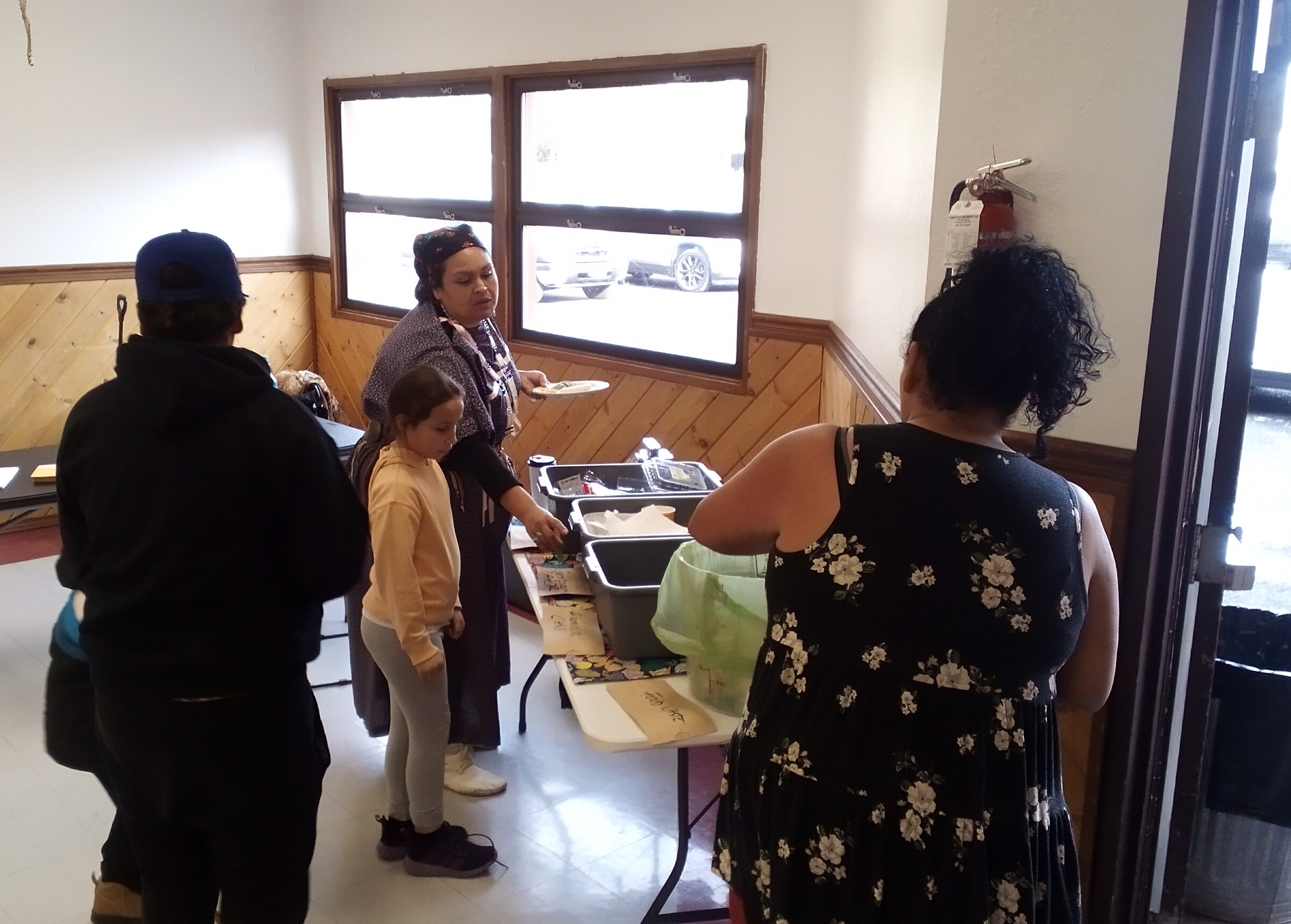 Several people stand around a table with multiple gray bins on it that each contain a different category of waste; paper, tin and aluminum cans, and landfill can be seen written on cards in front of each bin.