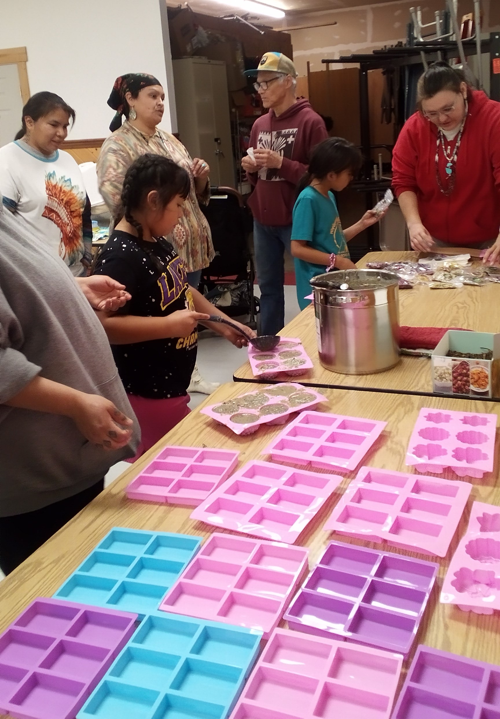 Several people stand in front of a Longhouse table covered with brightly colored silicon molds and a tall cooking pot with soap base as DeArcie explains the process of ladling out the soap base into the molds to dry.