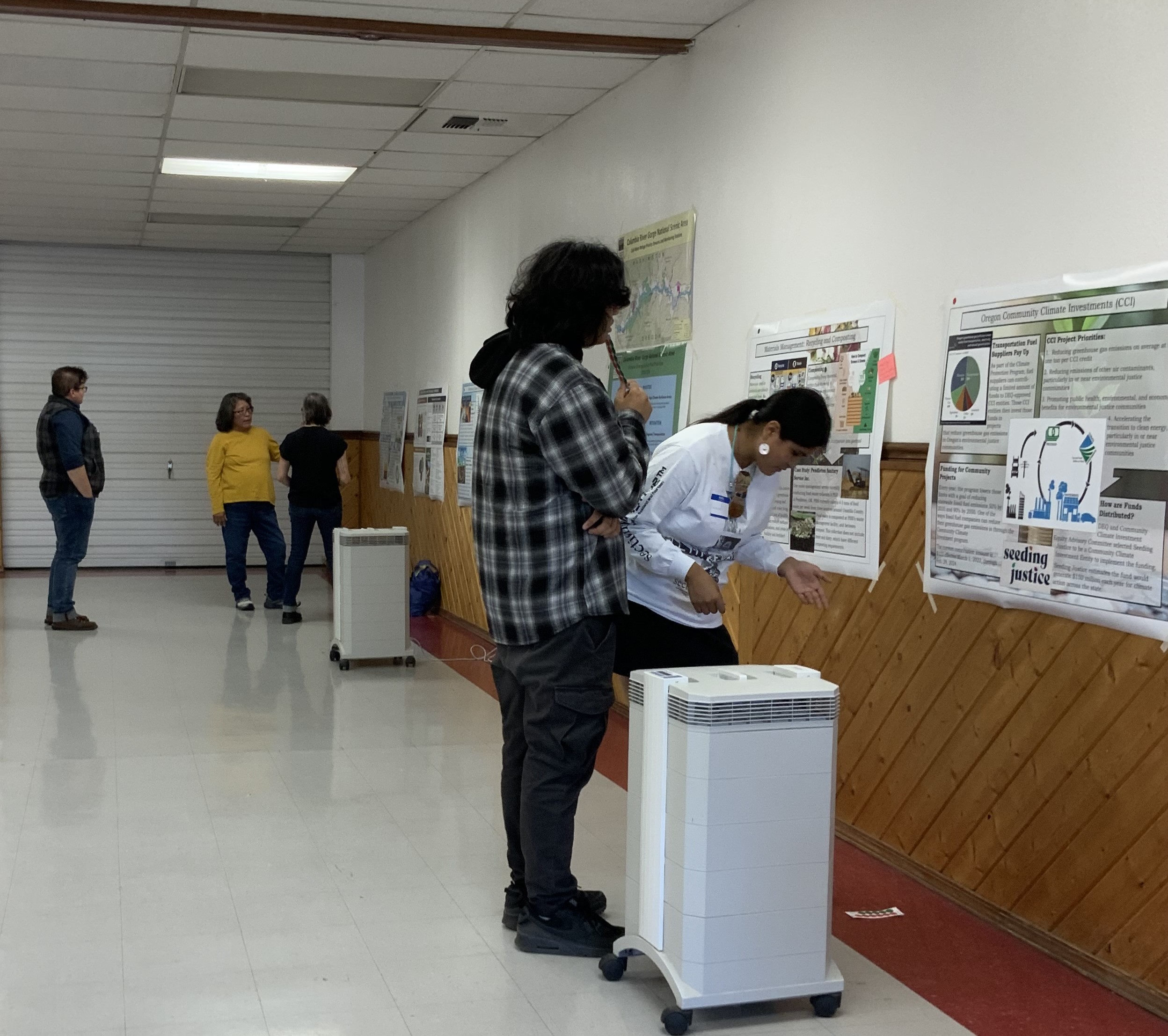 Two Tribal youth consider one particular carbon management strategy poster on the wall of the Longhouse annex, with three additional event participants discussing another poster in the background. Two high quality air purifier units are visible in the room.