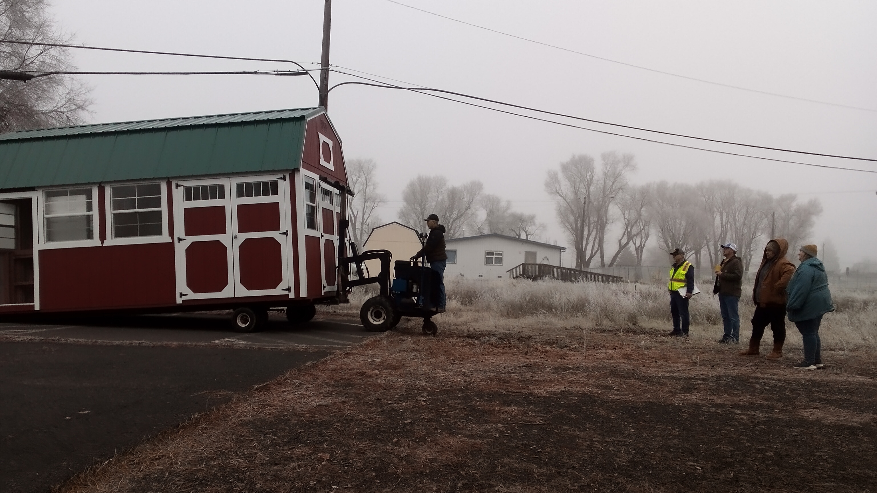 4 people dressed in winter clothing stand aside and watch a 5th person operating a fork lift lower a red utility shed onto pavement.