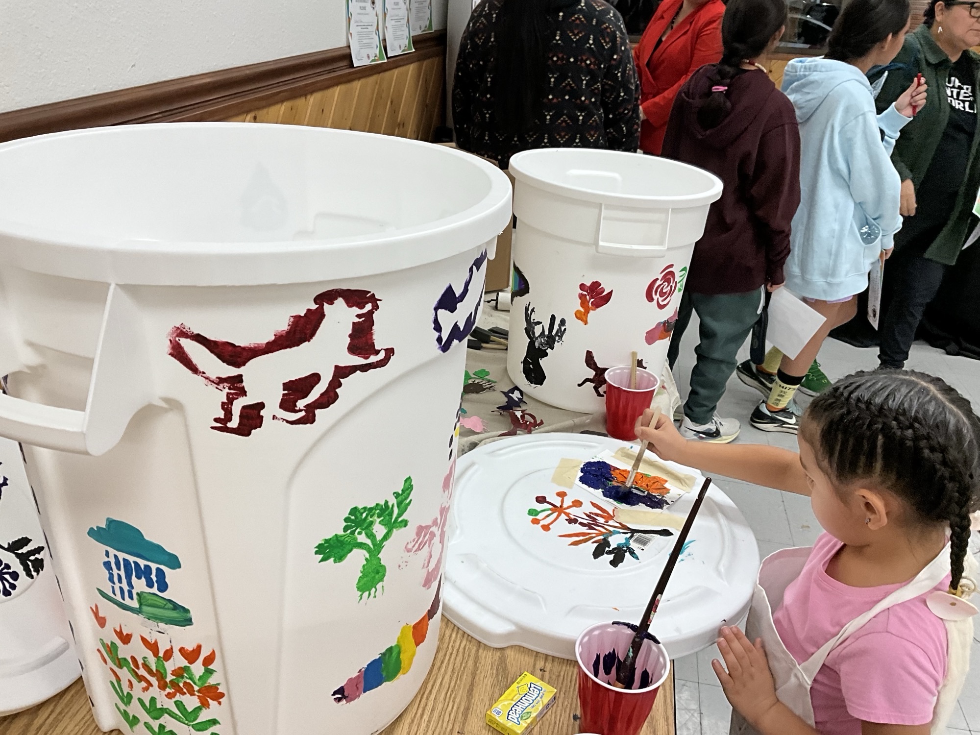 A Tribal young person in a pink t-shirt and braids paints a white trash bin with colorful shapes as others gather in the background.