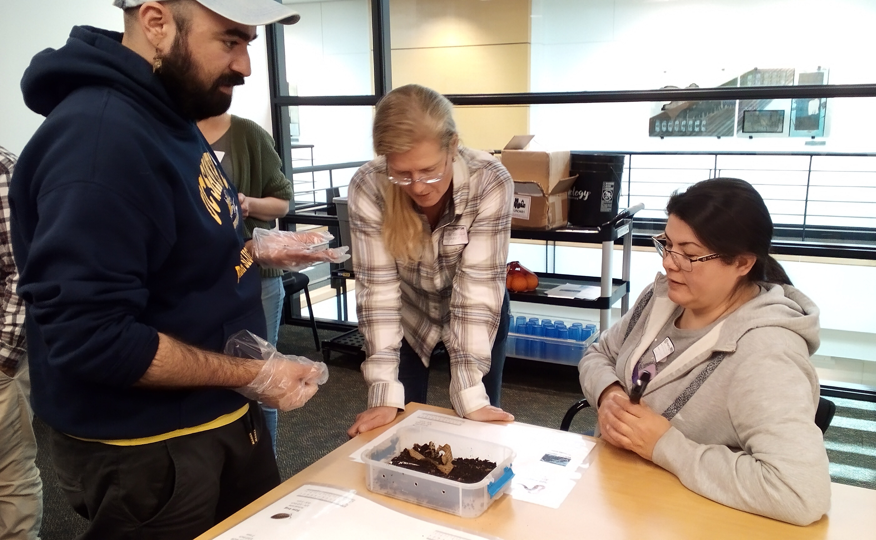 Three people are gathered at a table looking down at a plasticware container with dark soil inside of it.