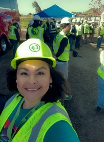 DeArcie smiles at the camera in a selfie wearing a bright yellow hard hat and safety vest, in the background is a group of other people also wearing hard hats and vests.