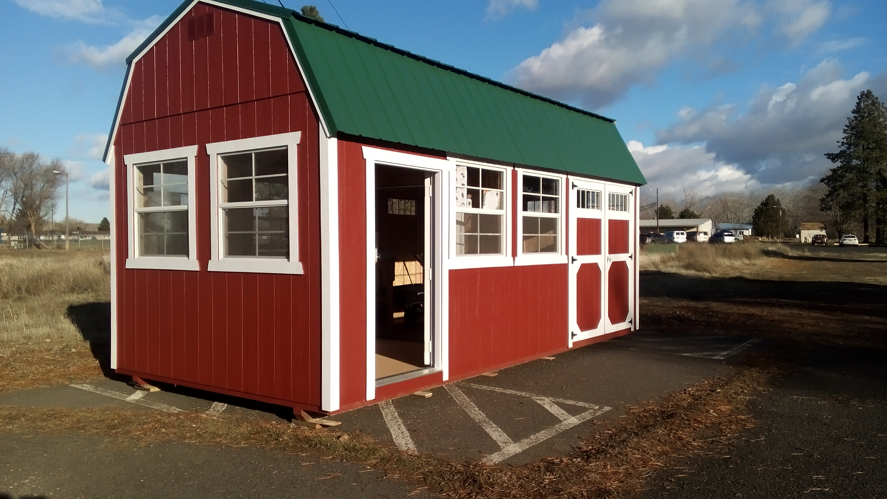 A red utility shed with a green metal roof and white trim sits on a pavement slab in front of a field of overgrown vegetation. The sky behind it is blue with white clouds, and the sunlight appears like late afternoon in winter.