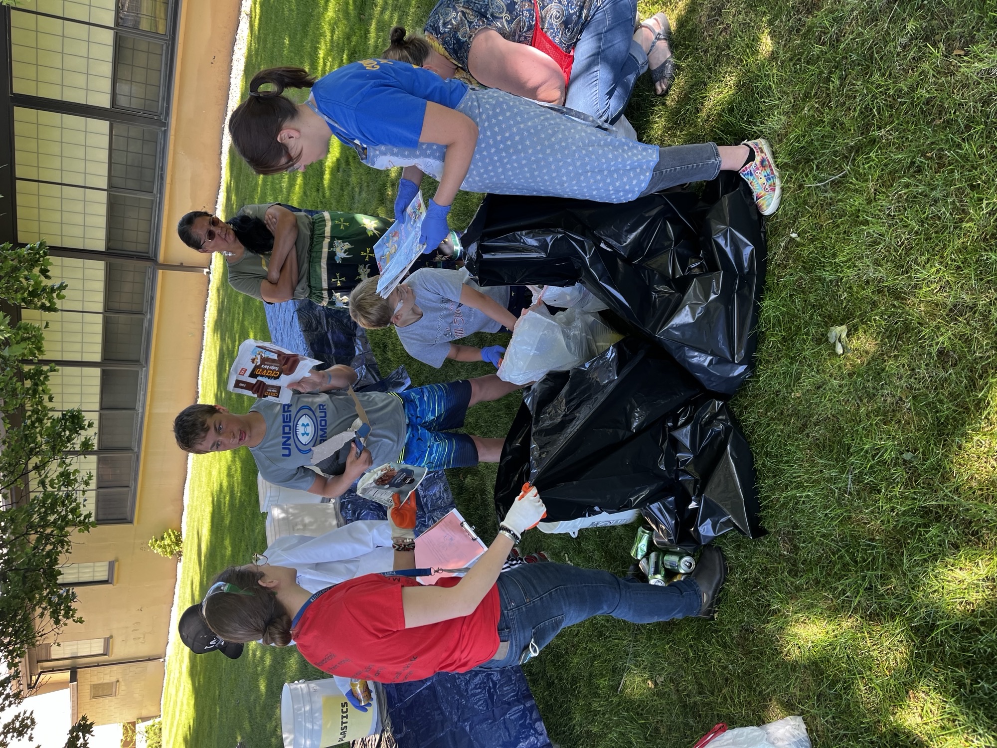 A group of students and teachers gathers around several large black trash bags outside in the shade. Students hold up and discuss different items from the trash.