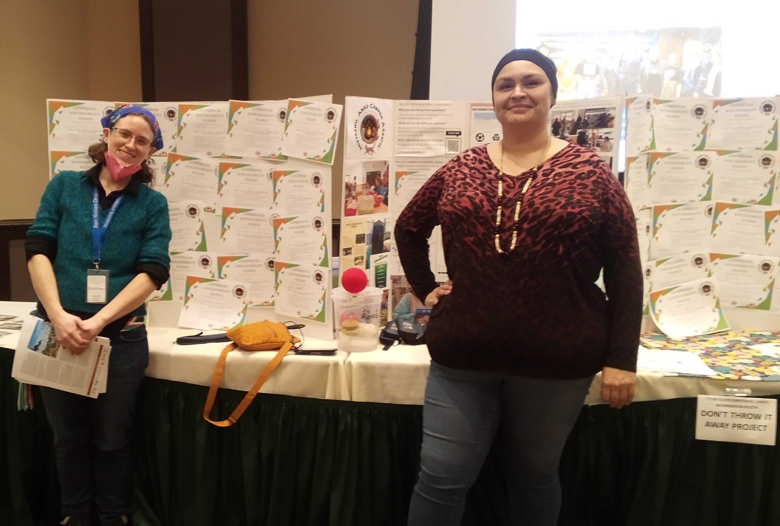 Two people stand in front of a table with a large cardboard trifold stands in the Wildhorse Rivers Event Center Cayuse Room, covered with colorful infographics, photos, and certificates.