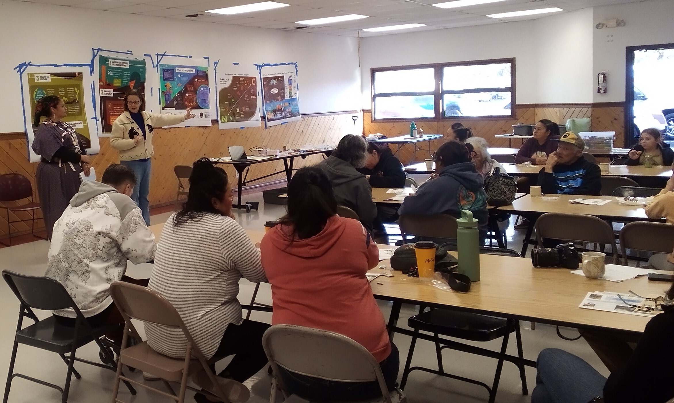 About 20 people sit in the brightly lit Longhouse annex room at long tables; two people stand in front of posters taped to the wall and explain project elements to the audience.