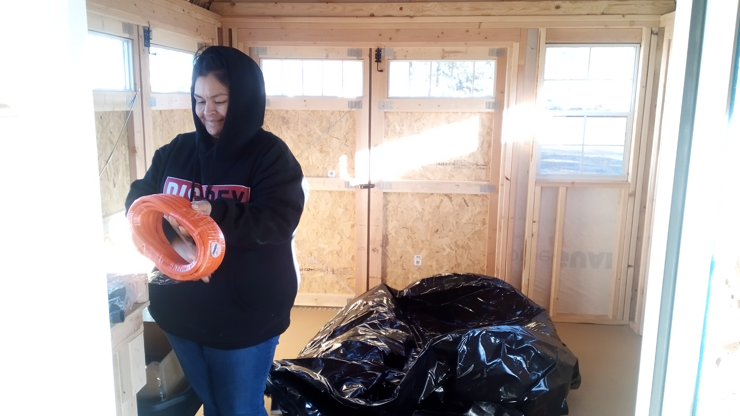 A person in a black hoodie with the hood up over their head stands in a brightly sunlight utility shed holding a coil of orange tubing. Around them is a large black plastic bag and tools and other pieces of equipment on the shed floor and shelves.