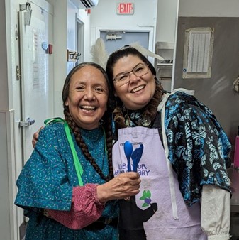 Two women smile in blue wing dresses widely in the Longhouse kitchen; the woman on the left holds up blue bioplastic utensils from the kit to demonstrate she brought them for Feast.