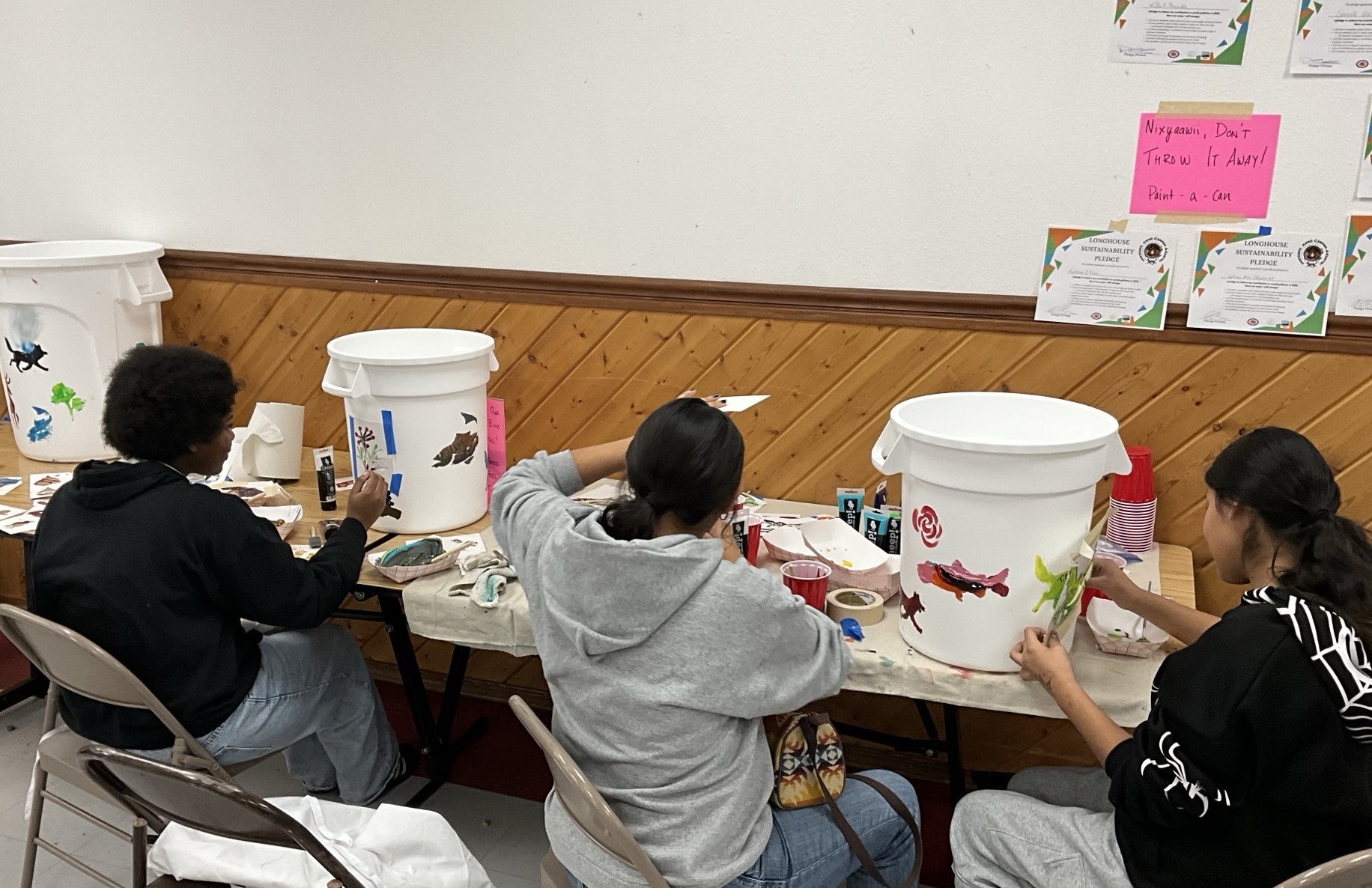 Three youth sit at a Longhouse table and paint two white trash bins with colorful shapes.