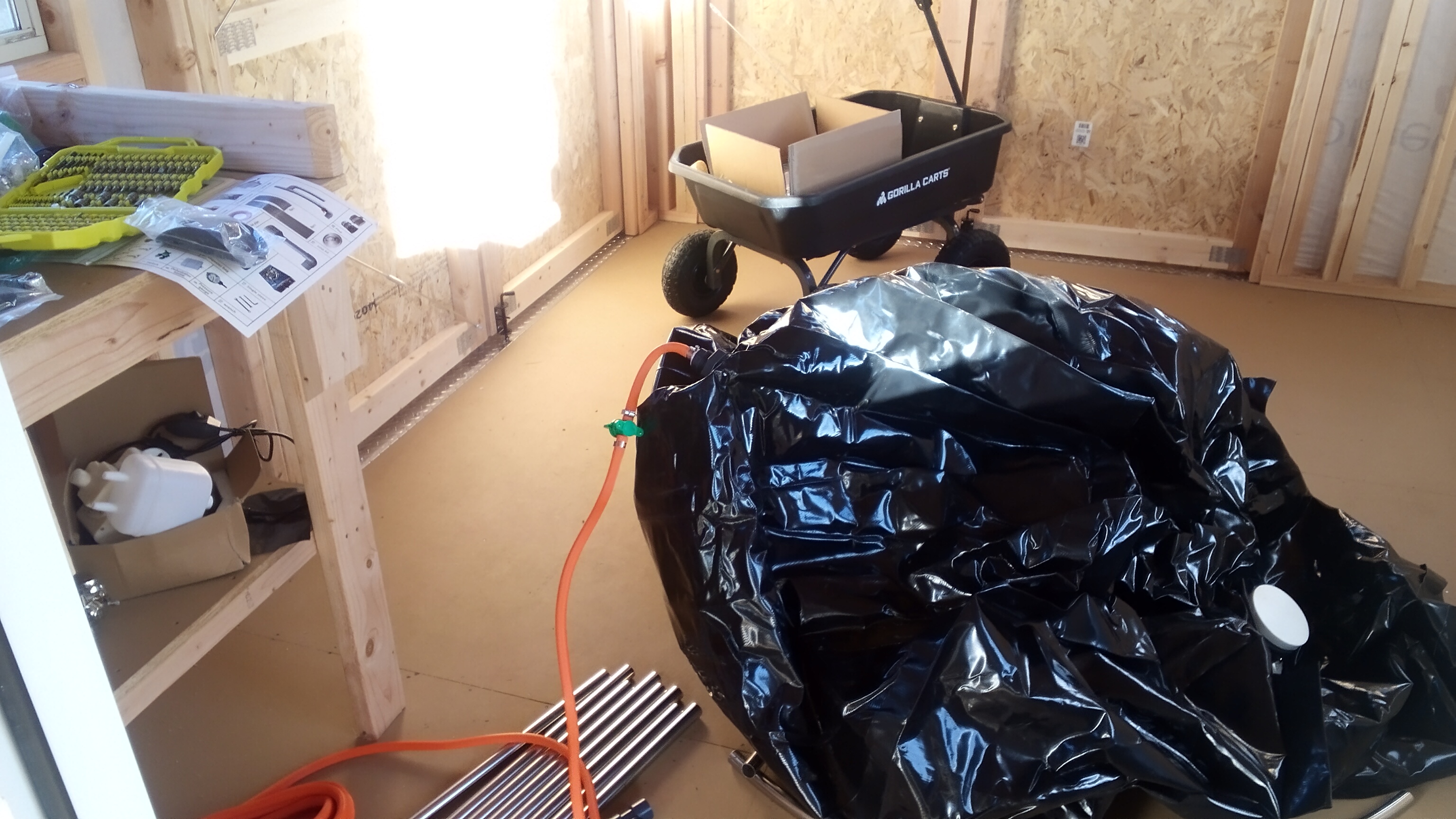 A large black plastic bag sits in the center of the interior of a utility shed with sunlight coming in through windows on all sides of the walls. Metal pipes and other parts are on the floor and shelves of the shed, and a sturdy four-wheeled pull behind cart has a carboard box inside of it.