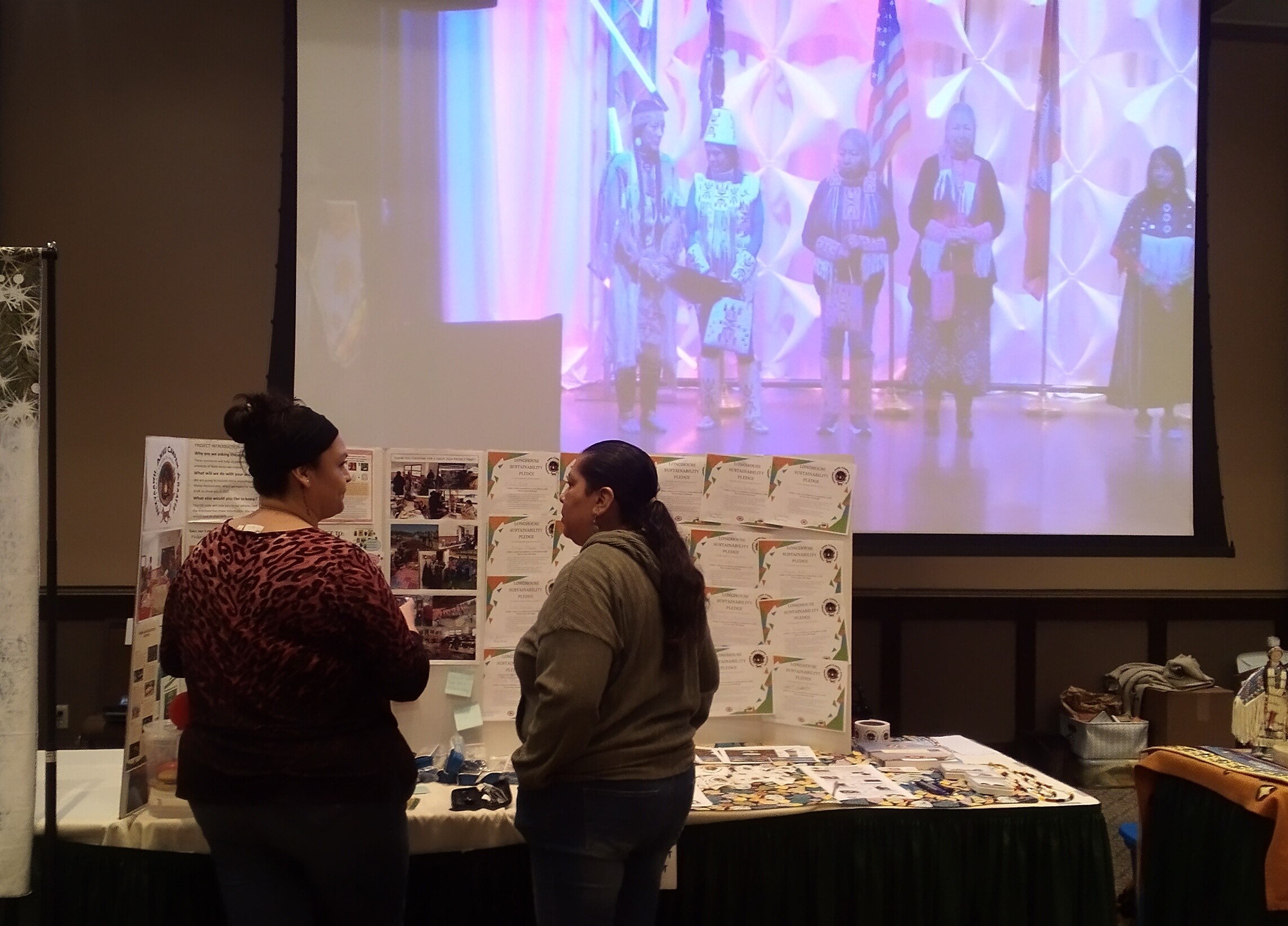 Two people stand in front of a table with several large cardboard trifolds covered with colorful photos, infographics, and certificates. In the background a large screen shows many people on Wildhorse Rivers Event Center stage on screen.