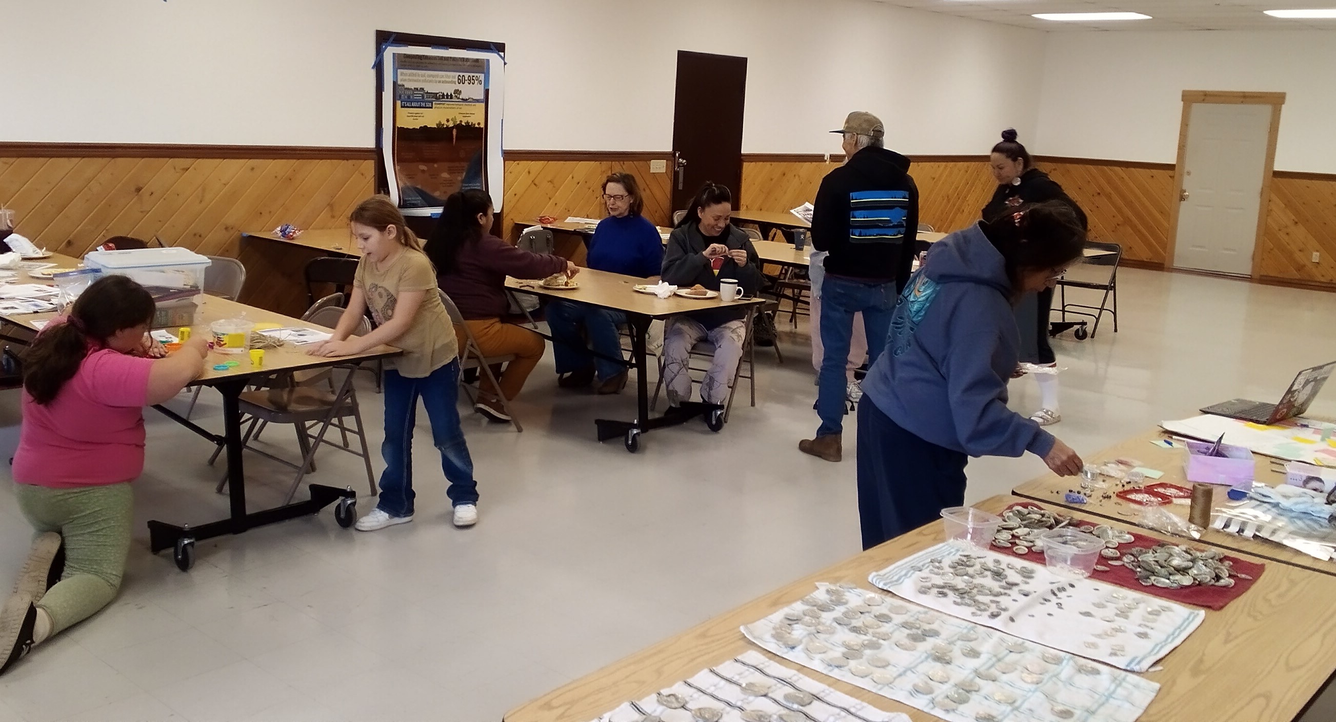 A group of people laugh and smile as sit at long tables in the Longhouse annex room after they get refreshments and earring making materials at the start of the event.