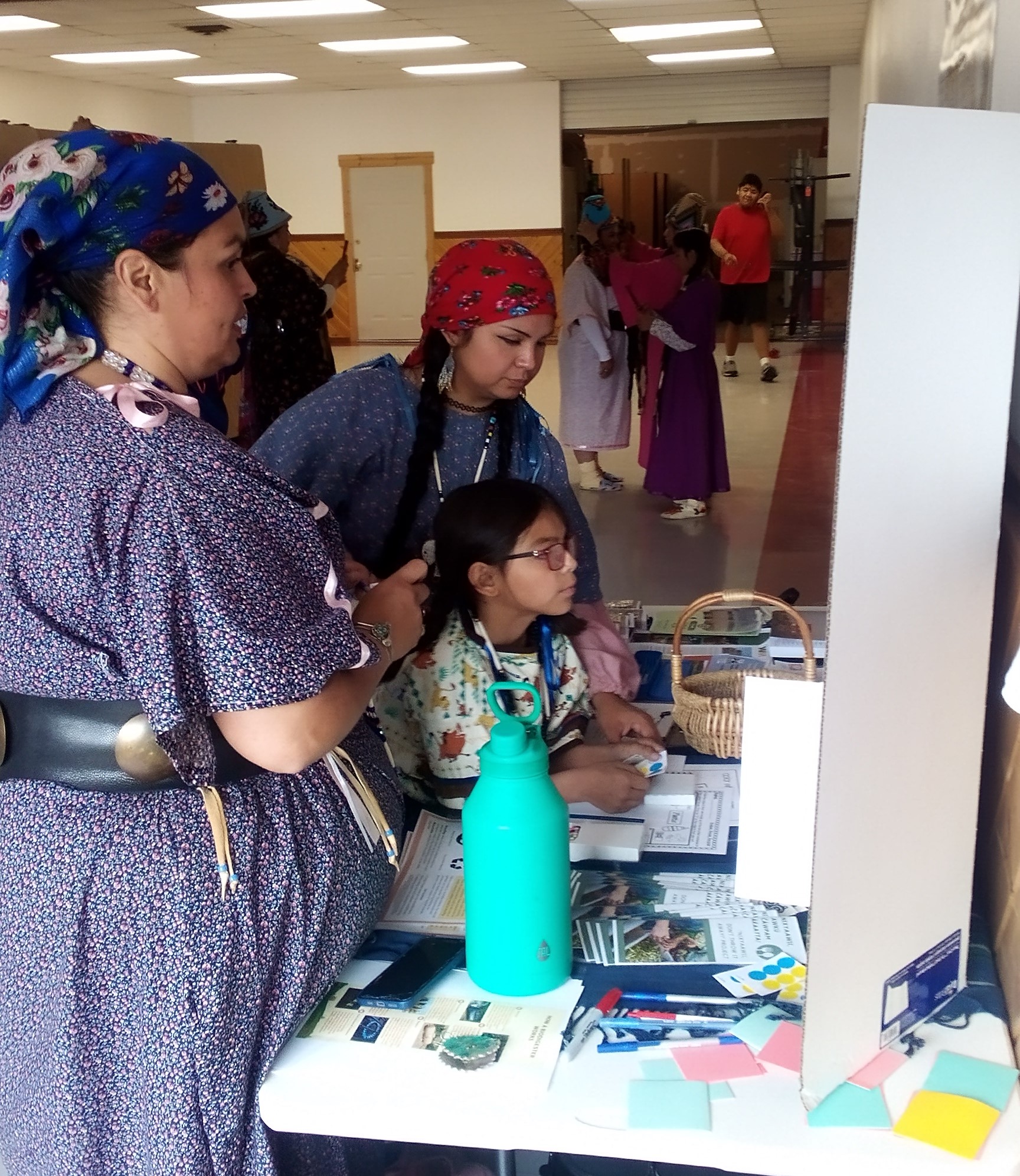 Several people including a youth considers images and text on a white poster board as other kids entertain themselves in the background of the Longhouse annex.