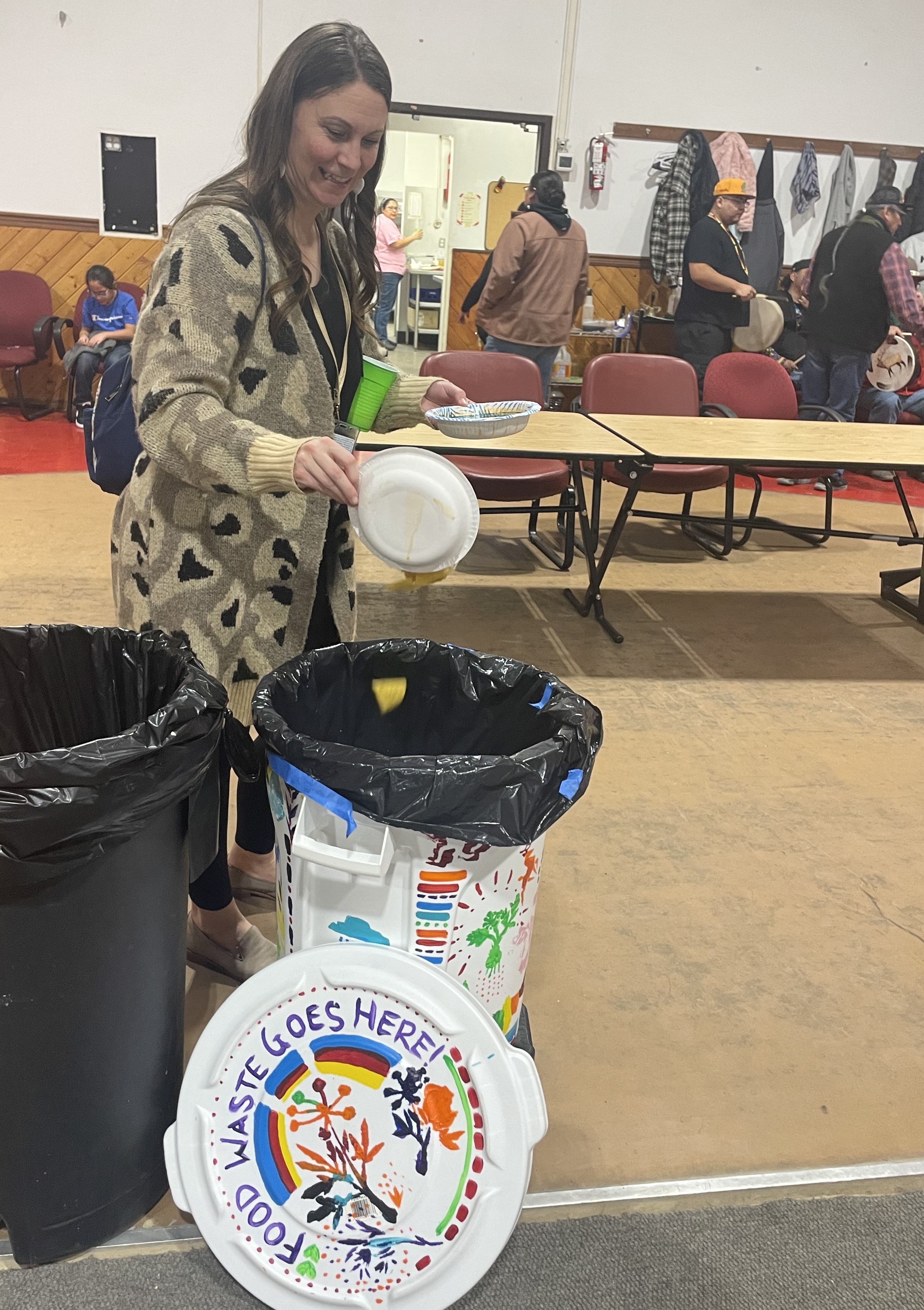 A Tribal family member smiles as she pours her left over food into the painted food waste collection bin a the Longhouse. The lid of the bin can be seen on the floor in front of the bin, and it reads “food waste goes here!”