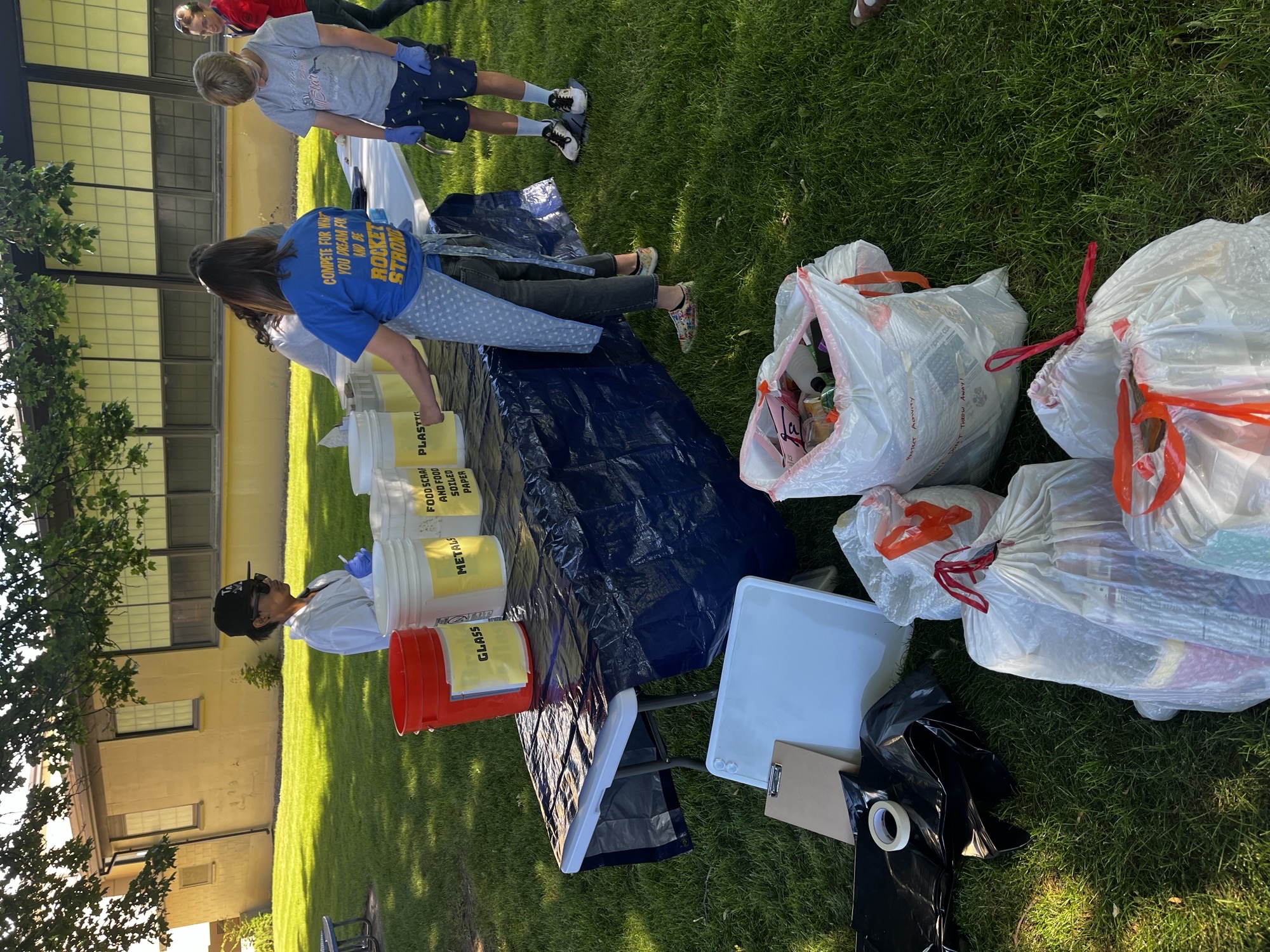 A group of students and teachers gathers around tables covered with blue tarps as they set up buckets for each waste category outside in the shade. Buckets are labeled with “Glass,” “Paper,” and other category names