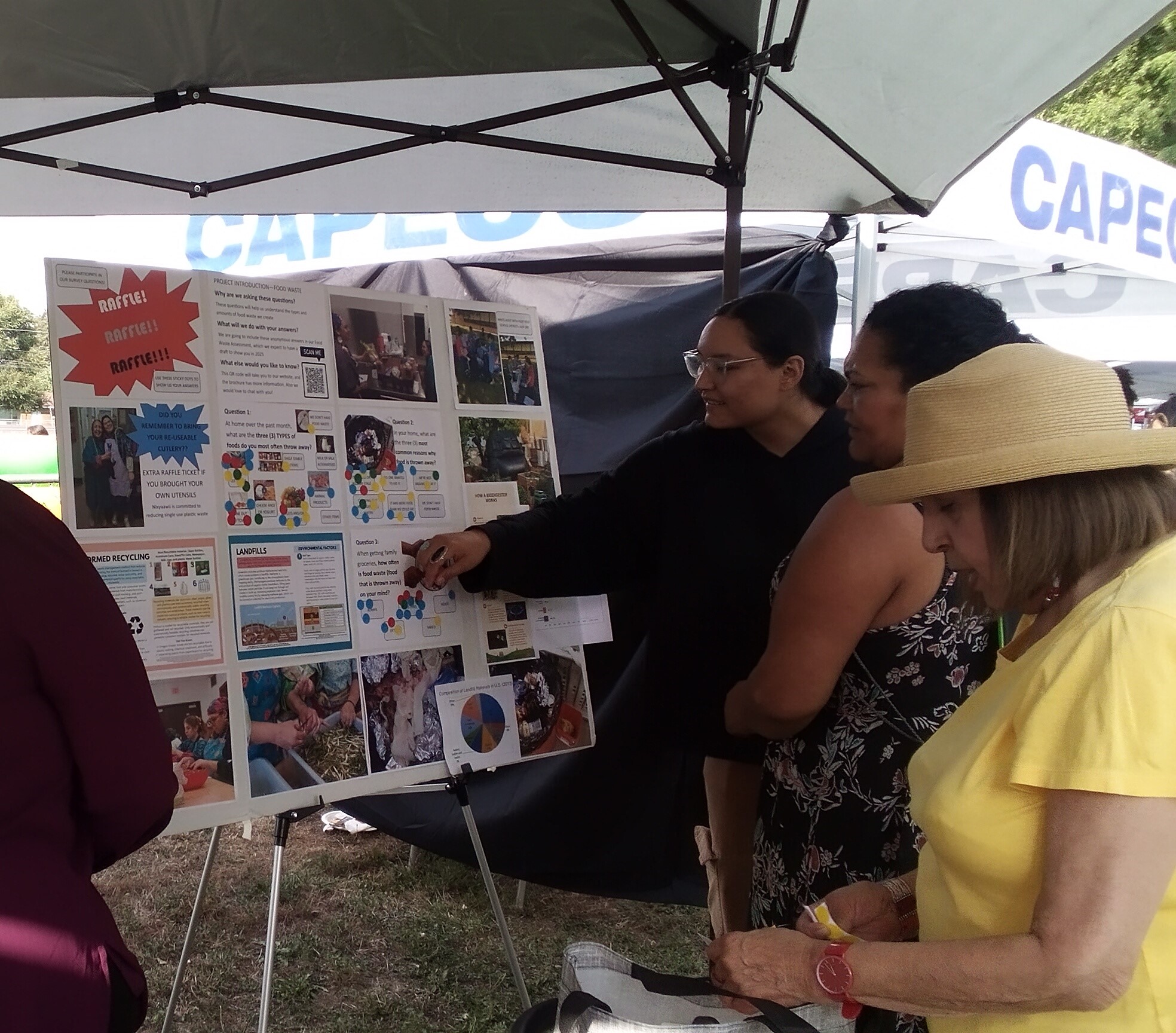 Three people gather around a display board considering food waste survey questions under a canopy in the shade.