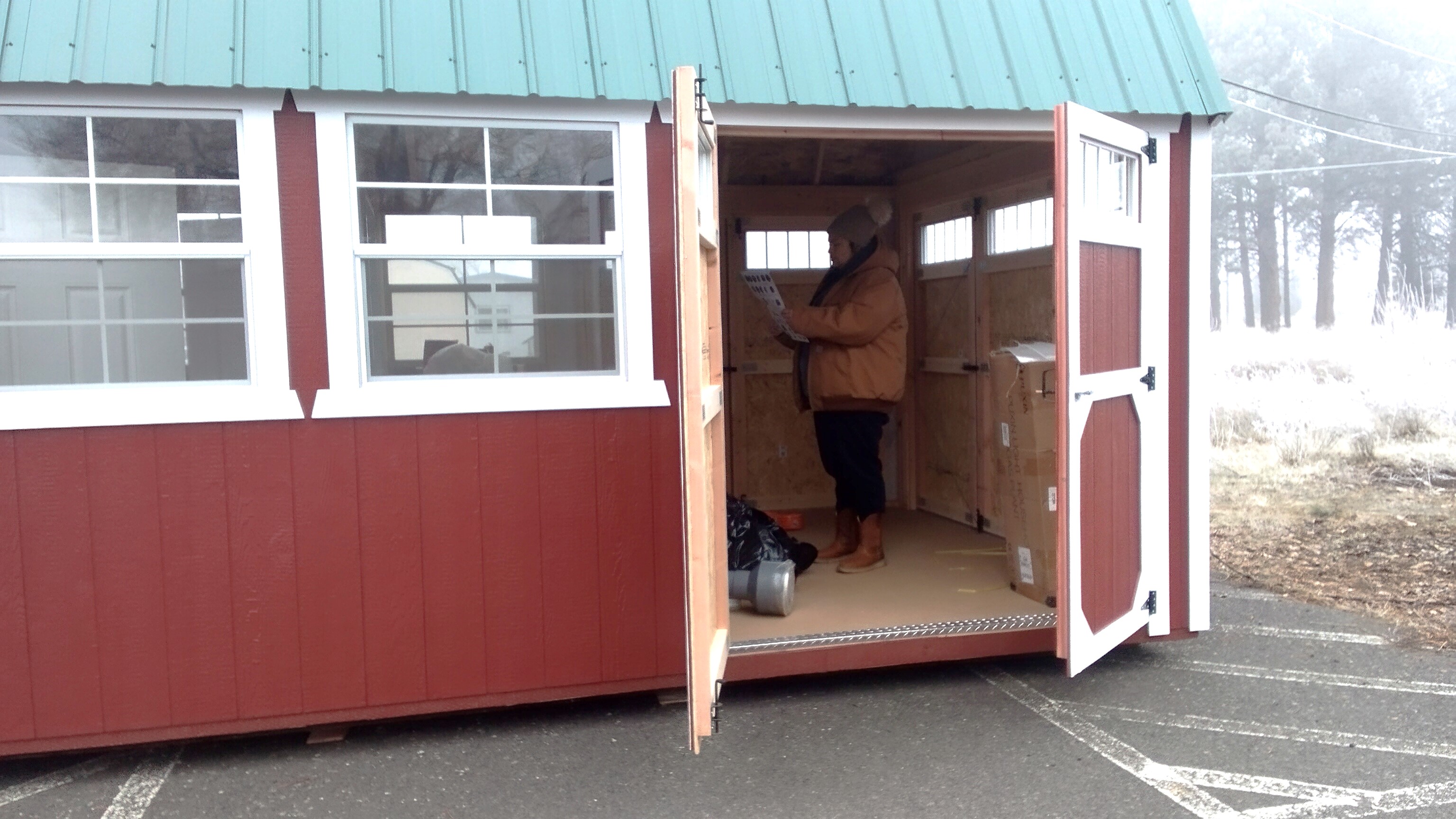 A person dressed in winter clothing stands inside a red utility shed and looks at a piece of paper, plumbing parts and a large plastic bag that will be the digester sits on the floor of the shed around them.