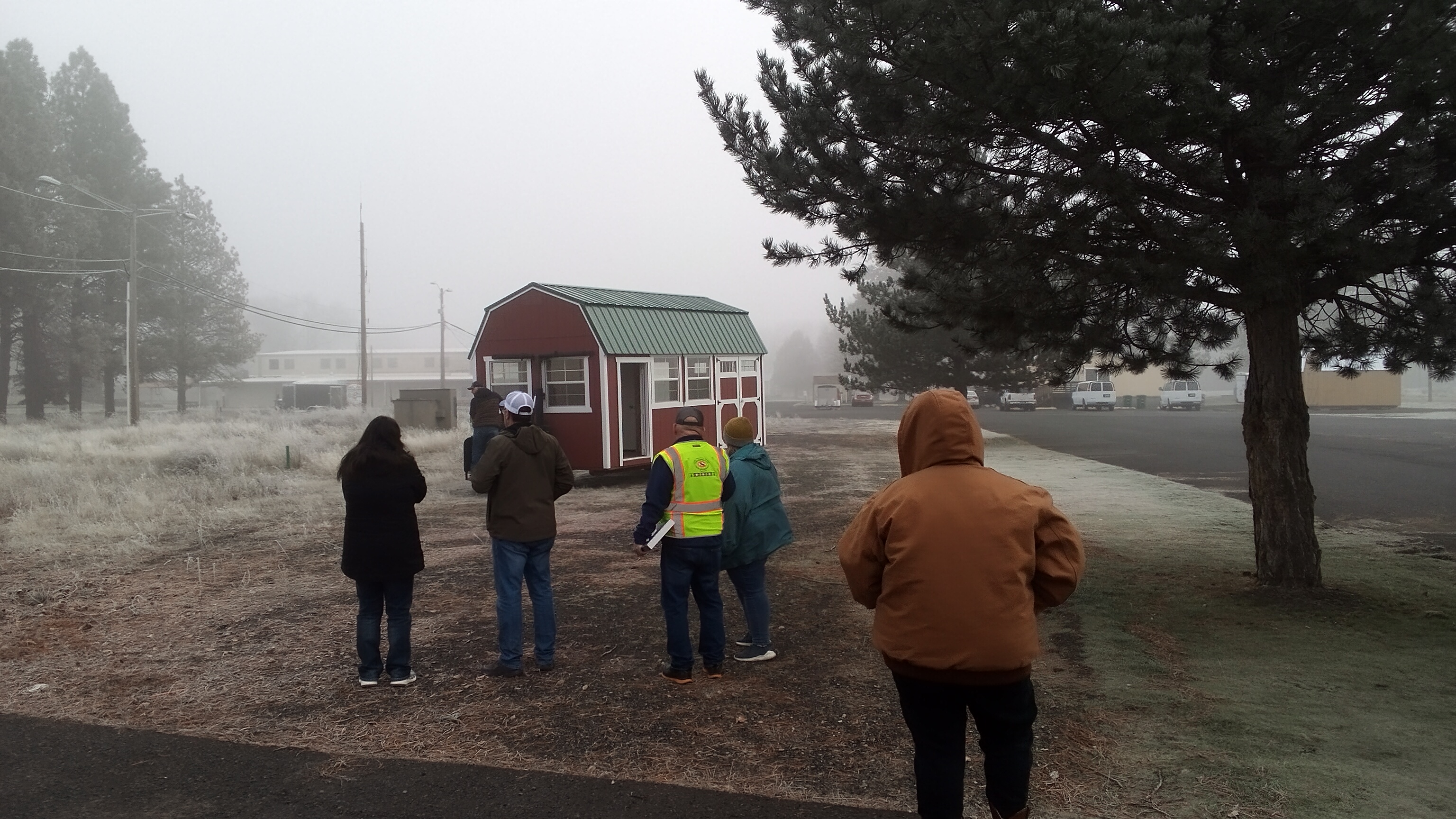 5 people dressed in winter clothing stand in a field as they observe a red utility shed approach its installation site.