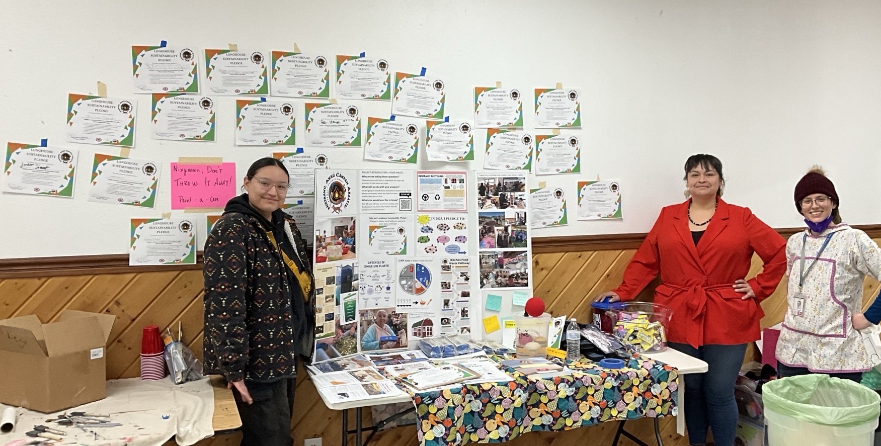 Three people stand beside a large cardboard trifold display and numerous colorful certificates taped to the wall above them.