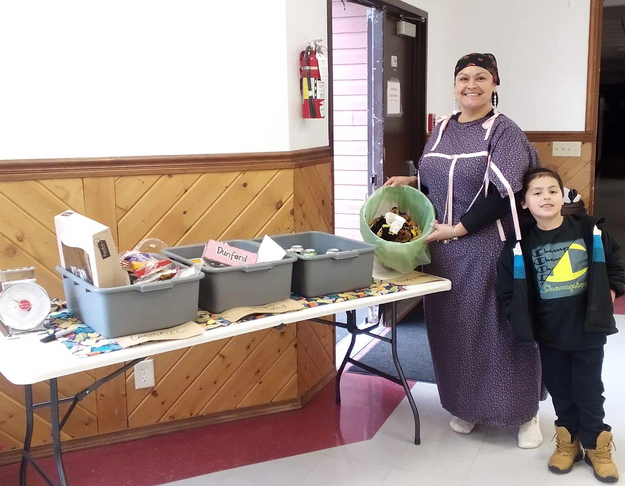 A woman and child stand together smiling as one shows off a bucket of food waste collected. On the table next to them is a table with several gray bins each with different kinds of waste.