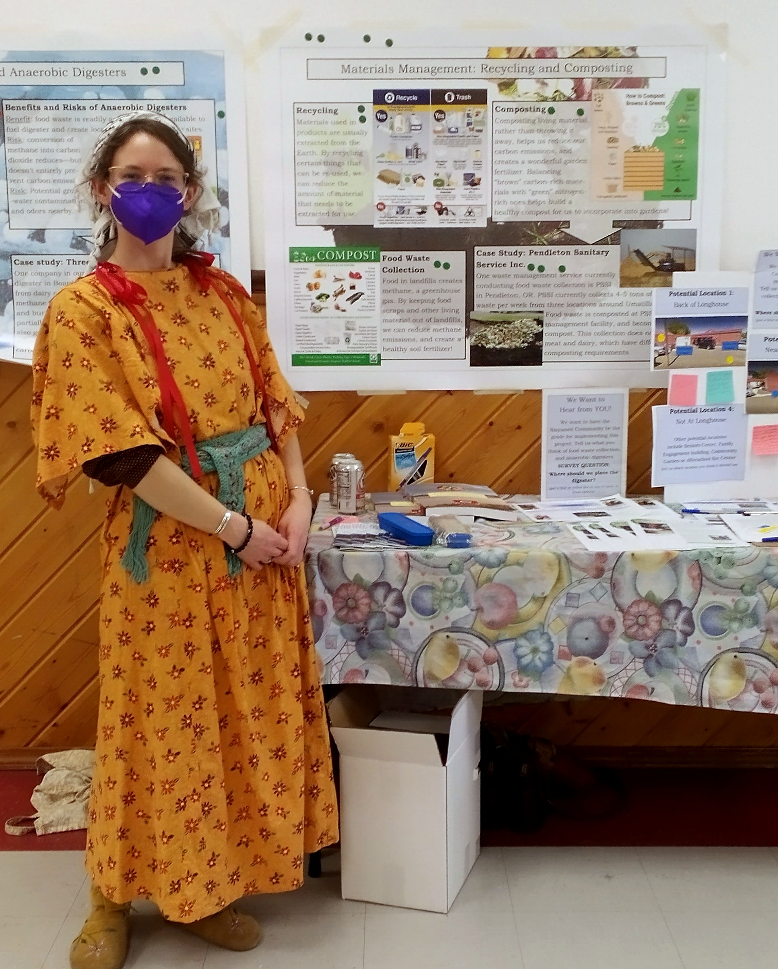 A person stands at the project outreach booth in a wing dress, glasses, and KN95 mask in front of informational posters with images and text.